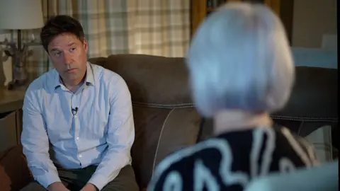 A blurry image of the back of Maria's head and white hair, as she talks to the reporter, who is wearing a blue shirt with a small microphone attached.