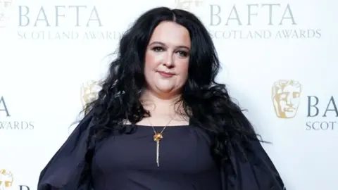 Ashley Storrie poses for the cameras at the Bafta Scotland Awards, wearing a black dress with puffed sleeves and an gold necklace.