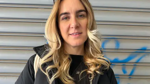 An image of Olivia Graham stood outside corrugated metal shutters as she worked on the podcast. She is wearing a black jacket and top with her blonde/brown hair down in curls around her shoulders. 