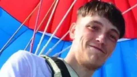 Family Photo Liam Woolford, smiling at the camera. He has short dark hair with fringe covering his forehead, and is wearing a white t-shirt. Red and blue fabric can be seen behind him with a number of poles holding it up.