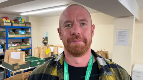 Ben looking into the camera and smiling slightly; he's wearing a black tshirt with a yellow, navy, and white checked shirt over the top, and has a bright green Trussell Trust lanyard. He's stood in front of tables full of food parcels, which are packed into brown paper bags.