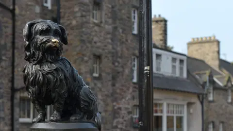 The Greyfriars Bobby statue in Edinburgh - a bronze terrier-type dog with a shiny nose where passers-by have stroked it. In the background are some historical terraced stone buildings.