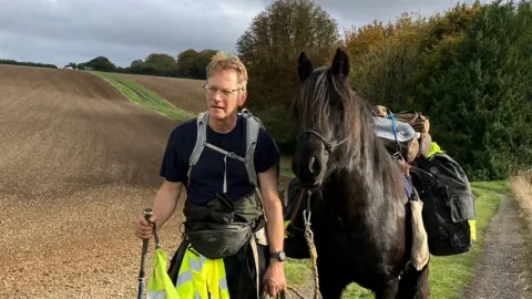 Roger Sewill during a trek in a field next to Scarlet. She is carrying his bags.