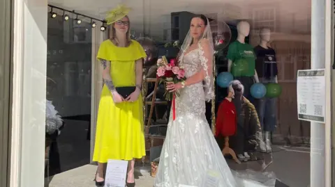 Alec W G Hall Ltd Nat wears a canary yellow dress and matching hat and Hattie has full bridal wear on complete with bouquet of pink and red roses.