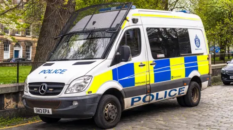 A police van with a riot shield attached to the windscreen. It is parked on a cobbled street.