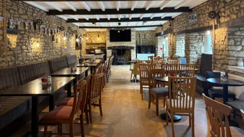 The inside of a pub with brick walls and a fire place and rows of empty tables and chairs.
