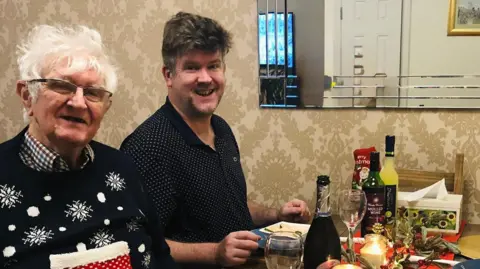 David Williams A man with grey hair and classes sits next to a younger man at a dinner table with food and drink.