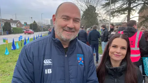 A man with a small amount of short grey hair and a grey beard stands next to a woman with long, straight reddish brown hair. He is wearing a blue coat with the Tewkesbury Academy logo on it, along with others, and she is wearing a black coat.