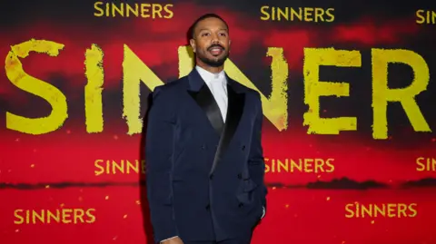 Reuters Michael B Jordan in a dark blue tuxedo standing in front of a red backdrop with Sinners in large yellow letters