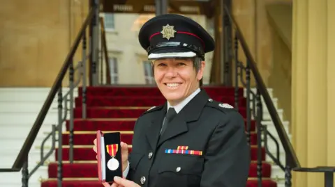 Suffolk County Council Ms Hammond smiles as she holds her Queen's Fire Service Medal. She wears a black uniform and a black hat. Stairs can be seen behind her.