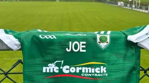 Green hurling top, with the name Joe written across the middle. Backdrop is a green hurling pitch