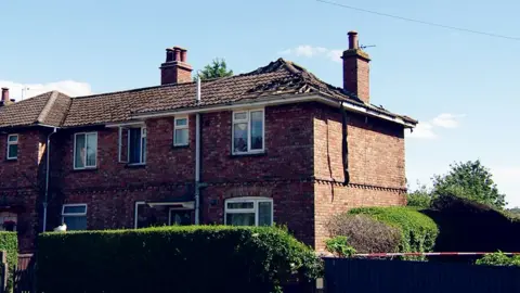 The fire-damaged house. There are damaged tiles on the roof and burn marks around the windows and door.