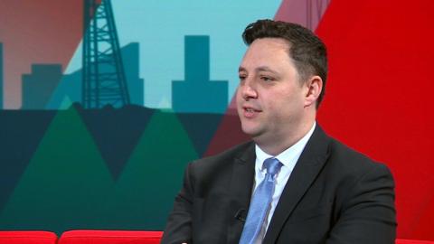 Ben Houchen sits in the red Politics North studio. He is wearing a black suit and a light blue tie.