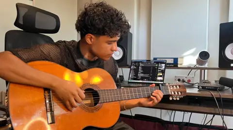 Kamal plays guitar in a studio surrounded by a laptop and speakers. He is wearing black trousers and wears a black top.