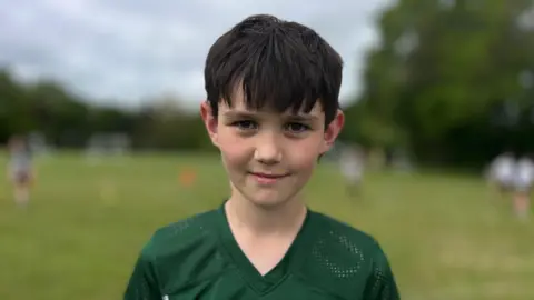 Martin Giles/BBC A boy wearing green sports kit. He has short dark hair and is standing outside on a grass pitch. He is looking directly at the camera and smiling.