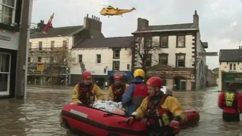 Three emergency workers wearing yellow dry suits with red helmets and life guards push a red inflatable boat along a partially submerged street. A yellow helicopter is in the air hovering behind them.