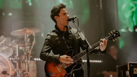 Stereophonics' Kelly Jones performing on stage. He is holding a guitar and singing into a microphone, wearing a leather jacket. 