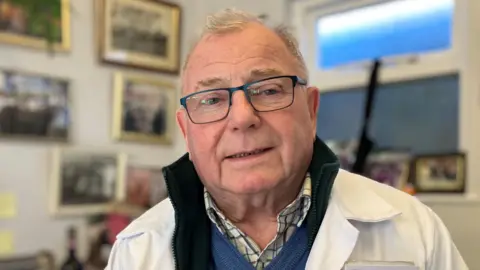 Lewis Adams/BBC John Coleman, who is wearing a white butcher's coat over a navy jumper and chequered shirt. He has glasses on and is smiling while sitting in his office, which has framed pictures on its wall.