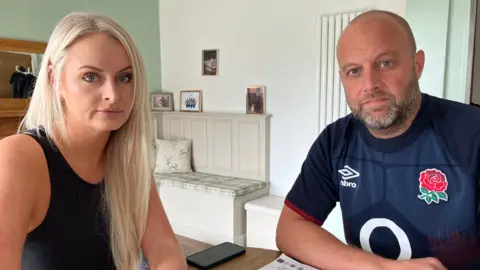 BBC Emma and James Richardson sit at a table in their smartly decorated home. Emma has long blond hair and is wearing a black sleeveless top. James has short fair hair and a neatly trimmed beard and is wearing a blue England rugby shirt. 