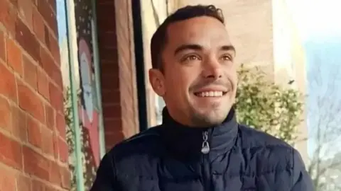 Family Handout A man with brown hair and brown eyes smiling in the sunshine whilst wearing a black puffer jacket that has a silver zip. 