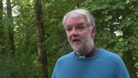 Reverend John Dixon stands in front of some trees. He is wearing a dog collar and a blur jumper. He has short grey hair and a beard.