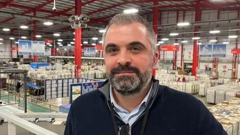 BBC/Phil Harrison Tom Bailey, plant manager at the Medway Mail Centre, pictured overlooking the centre floor. Tom is wearing a navy pullover jumper with grey collar, a black lanyard and a blue shirt underneath. He has glasses hooked onto the front of his jumper.