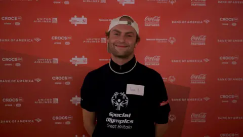 A young man with stubble and a white backwards baseball cap smiles. 