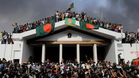 Getty Images Anti-government protestors display Bangladesh's national flag as they storm Prime Minister Sheikh Hasina's palace in Dhaka on August 5, 2024. Bangladesh army chief Waker-Uz-Zaman spent nearly four decades rising to the top of the military and said on August 5, he was "taking full responsibility" after Prime Minister Sheikh Hasina was ousted and fled