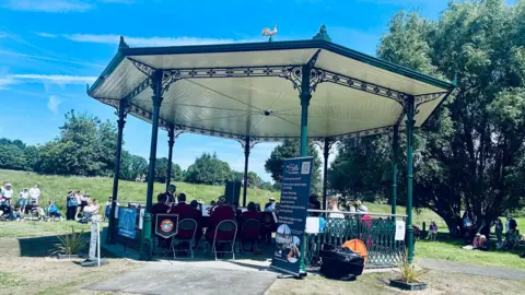 Claudia Sermbezis / BBC A bandstand in a sunny park with metal fencing and poles around the sides, a pale wood cladded roof and a golden cockerel on the top. A band is sitting on chairs and playing inside the bandstand, and there is a crowd on the grass around the edges.