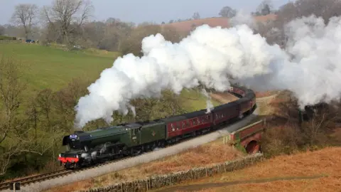 Severn Valley Railway/Ian Cawthorne An aerial view of the train, with steam coming from the locomotive and going from virtually the left to the right on the photo. There is grass on much of the left side of the image.