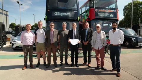 Eight travel representatives pose for a picture in front of two buses at the depot. Luke Marion in the centre holds up the letter.