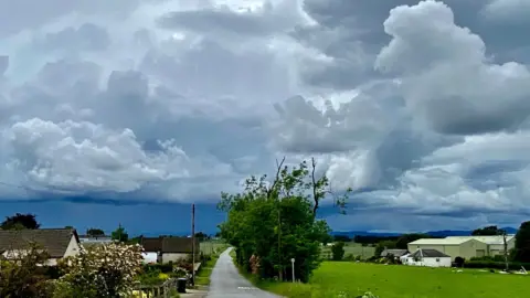 Lara/BBC Weather Watchers A rural scene of a road between fields. a tall hedge and houses. In the distance are large fluffy clouds with dark blue undersides.