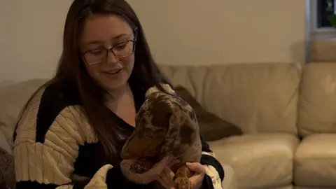 Kamelia Yotava holds a dachshund as she sits on a sofa. She wears a black and cream stripe jumper.