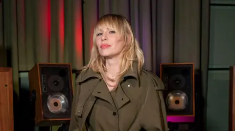 James Watkins/BBC Natasha Bedingfield wearing a green coat and standing the middle of two speakers. 