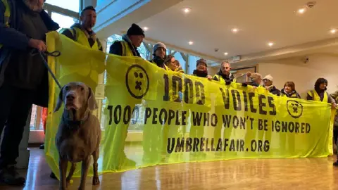 People standing behind a yellow 1,000 Voices banner in a large stone guildhall. Some are wearing hi-vis jackets. One has a brown dog on a lead.