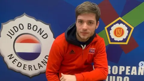 Rowan Kinsella Rowan wearing a red GB zipped top with an Adidas sponsor logo. He is in front of a board that reads Judo Bond, Netherlands and has the European Judo logo on it which is red, green and blue around a white flower and yellow pentagon.