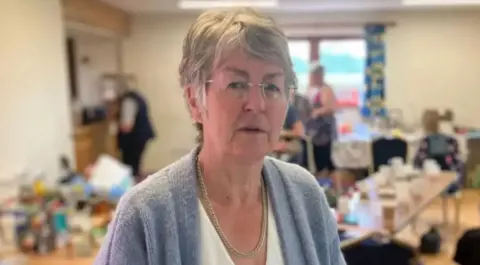 A woman with short grey hair and glasses with a white top and a pale blue cardigan in front of a blurred background 