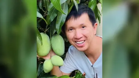 Thanh Phan smiling next to a mango tree with immature fruit