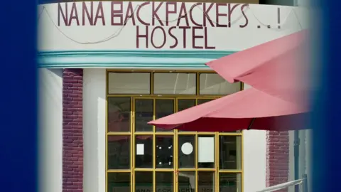 A view of the Nana Backpackers hostel taken between the bars of a blue fence. Two pink parasols stand in front of the building. 