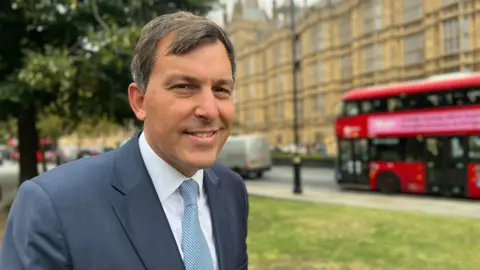 John Glen smiles at the camera in a suit and tie and is standing outside Westminster, London, with a big red bus behind him too