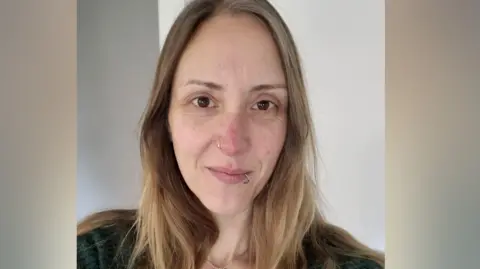 A woman with long blonde hair is looking at the camera. She has brown eyes and silver rings in her nose and lip. 