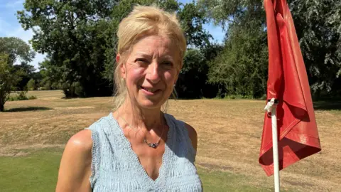 Jenny Kirk/BBC A woman with tied-back blonde hair is standing on a golf course next to a red flag. She is looking directly at the camera and is wearing a light blue vest top.