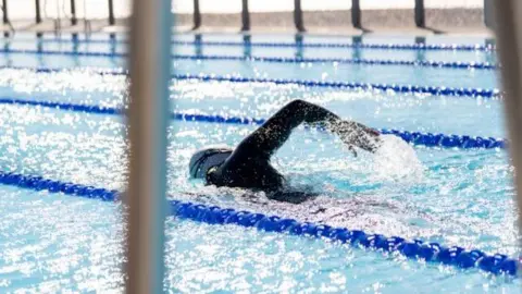 Supplied Someone swimming in an outdoor pool with plastic lane markers and the sun shining off the water. They are wearing a black wetsuit and a swimming cap.