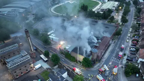 Eddie Mitchell A large plume of smoke is seen from above. It is pouring from the roof of a large supermarket. 