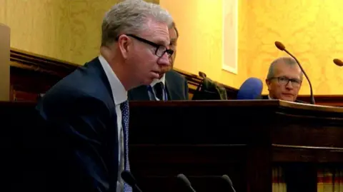 Allinson, a man with grey hair, he wears glasses and a suit as he stands up in Tynwald to speak.