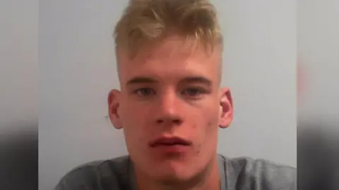 A police custody image of Jack Tomlinson, a white male with blonde hair wearing a grey t shirt.