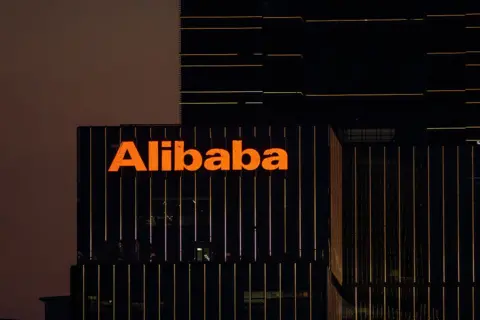 The Alibaba logo in orange lights is displayed on the exterior of an office that appears with black windows at dusk. 