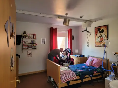 A bedroom with a door open that has Thea's name on it in cutout letters. A small picture of Thea is also on the door. The bed is made with blue covers that have a pattern of pink flamingoes and leaves. There is a bag for fluids hanging on a pole beside the bed. Shelves with books and toys on white walls. Dad Mark and nine year old Thea who is in her wheelchair are behind the bed. A picture of Mickey Mouse is on the wall above the bed.