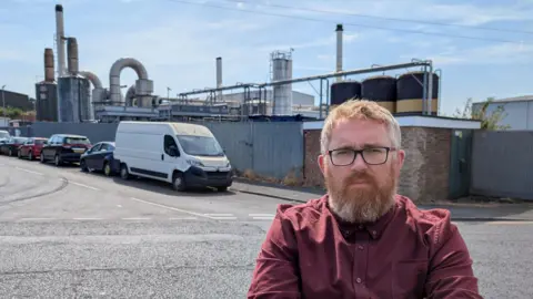 Matt Edwards/submitted A man stands in front of a factory plant, which has huge steel pipes and chimneys. The main has ginger hair and beard, and wears dark glasses and a maroon shirt. He has a serious look on his face.