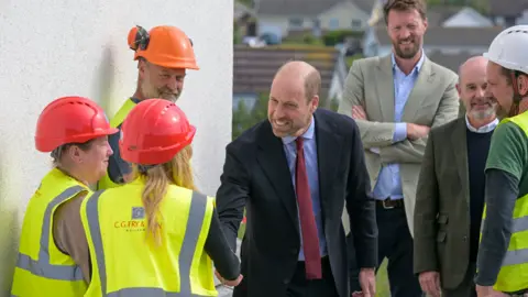 Prince William in a suit and red tie shaking hands with people in yellow hi-vis jackets. He is smiling as are those around him.